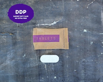 May include: A rectangular brown paper packet with a purple horizontal band featuring the white text TABLETS and the vertical text Multipurpose Cleaner, accompanied by a single white, oval-shaped, speckled cleaning tablet positioned below it.
