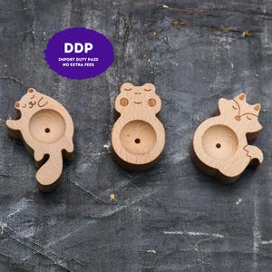 May include: Three light-colored wooden animal-shaped holders with circular indentations in the center. The left figure resembles a dog, the middle a frog, and the right a fox. Each piece features engraved facial details and a small hole in the center.