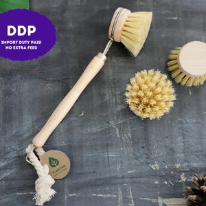 May include: A dish brush with a light wood handle and a round, stiff-bristled head, accompanied by two additional round replacement brush heads. A small, circular brown tag with a green leaf logo and the text PLANTISH is attached.