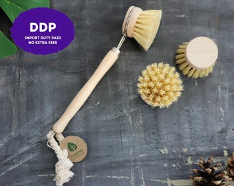 May include: A dish brush with a light wood handle and a round, stiff-bristled head, accompanied by two additional round replacement brush heads. A small, circular brown tag with a green leaf logo and the text PLANTISH is attached.