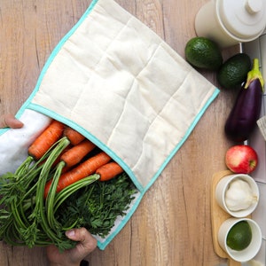 May include: A white fabric food storage bag with a turquoise trim, holding a bunch of fresh carrots with green tops. The bag is lying on a wooden surface.
