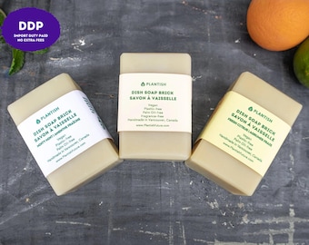 Puede incluir: Tres bloques rectangulares de jabón para platos de color beige claro, cada uno envuelto en una etiqueta de papel con texto verde. Las etiquetas dicen Plantish Dish Soap Brick, Savon à Vaisselle, e incluyen características como vegano y sin plástico.