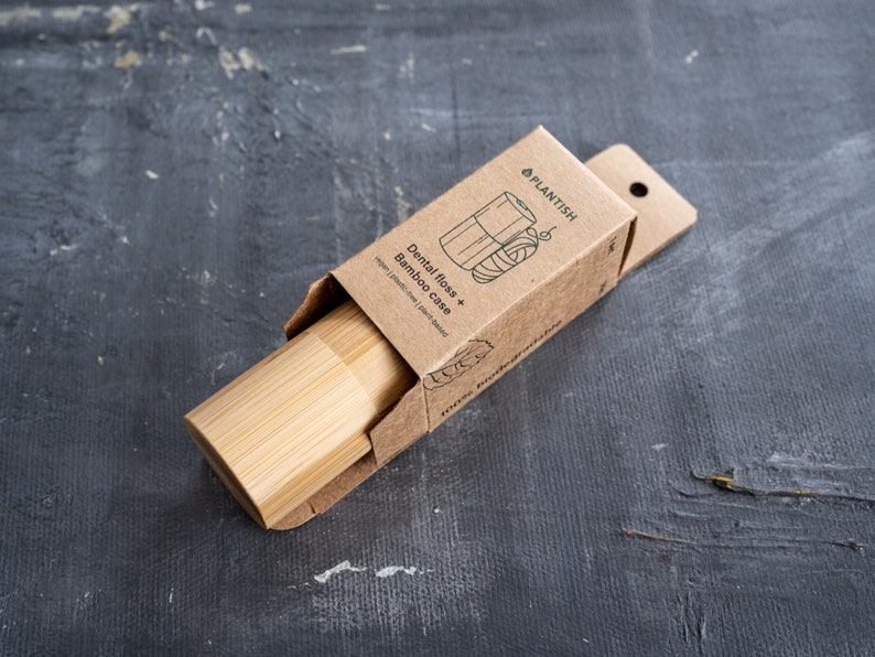 May include: A cylindrical bamboo dental floss case partially inside a brown cardboard box. The packaging features the text PLANTISH, Dental floss + Bamboo case, vegan | plastic-free | plant-based, and 100% biodegradable with a line art illustration.