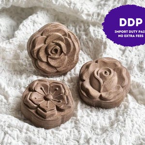 May include: Three circular brown wooden objects with carved floral designs. Two objects feature a layered rose pattern, while the third displays a five-petaled flower with a central stamen. The wood has a natural grain and a matte finish.