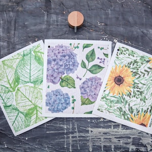 May include: Three decorative cleaning cloths with floral and leaf designs. One cloth features green leaves, another has purple hydrangeas, and the third displays yellow sunflowers. Each cloth has a small hole for hanging.