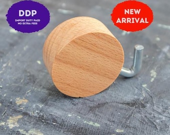 May include: A circular light brown wooden wall hook featuring a smooth, natural grain finish and a single silver-colored metal L-shaped hook extending from the side. The object is presented against a dark, textured background.