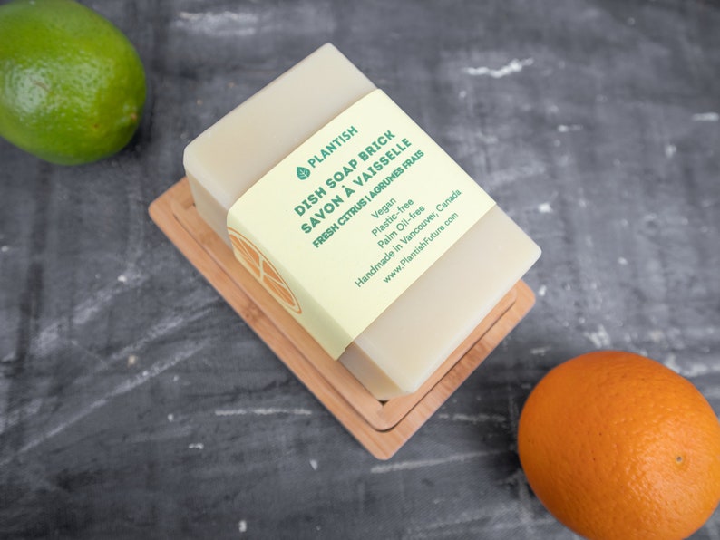 Pode incluir: Uma barra de sab&atilde;o para lavar lou&ccedil;a PLANTISH em um prato de madeira. O sab&atilde;o &eacute; etiquetado como "DISH SOAP BRICK SAVON &Agrave; VAISSELLE FRESH CITRUS | AGRUMES FRAIS" e lista os ingredientes como veganos, livres de pl&aacute;stico e livres de &oacute;leo de palma. O sab&atilde;o &eacute; feito &agrave; m&atilde;o em Vancouver, Canad&aacute;. O r&oacute;tulo tamb&eacute;m inclui o endere&ccedil;o do site: www.PlantishFuture.com.