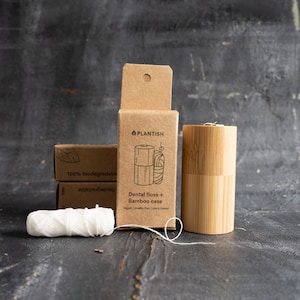 May include: A cylindrical bamboo dental floss case, a brown cardboard box labelled Plantish Dental floss + Bamboo case, and a spool of white dental floss. The box features a line drawing of the case and floss, with text stating vegan, plastic-free, and plant-based.
