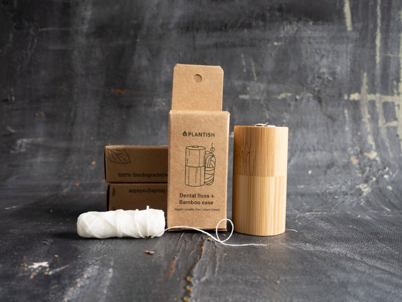 May include: A cylindrical bamboo dental floss case, a brown cardboard box labelled Plantish Dental floss + Bamboo case, and a spool of white dental floss. The box features a line drawing of the case and floss, with text stating vegan, plastic-free, and plant-based.