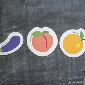 May include: Three round, white coasters with colourful fruit illustrations. The coasters feature a purple aubergine, a peach, and an orange. Each coaster has the word "Plantish" printed on it.