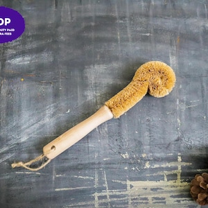 May include: A dish brush with a light-colored wooden handle and a curved, tan-colored fiber bristle head. The handle features a small hole at the end with a thin, natural-colored twine loop attached for hanging the cleaning tool.
