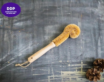 May include: A dish brush with a light-colored wooden handle and a curved, tan-colored fiber bristle head. The handle features a small hole at the end with a thin, natural-colored twine loop attached for hanging the cleaning tool.
