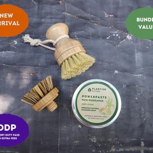 May include: Two round wooden dish brushes with light-colored natural bristles and a small white rope loop, alongside a circular metal tin of Plantish Future Powerpaste cleaning soap with a green label featuring lemon and plant illustrations.