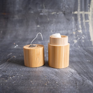 May include: A cylindrical bamboo dental floss container with a removable lid. The lid features a small metal cutter for the white floss, which is visible extending from the open container. The natural wood grain of the bamboo is clearly displayed.