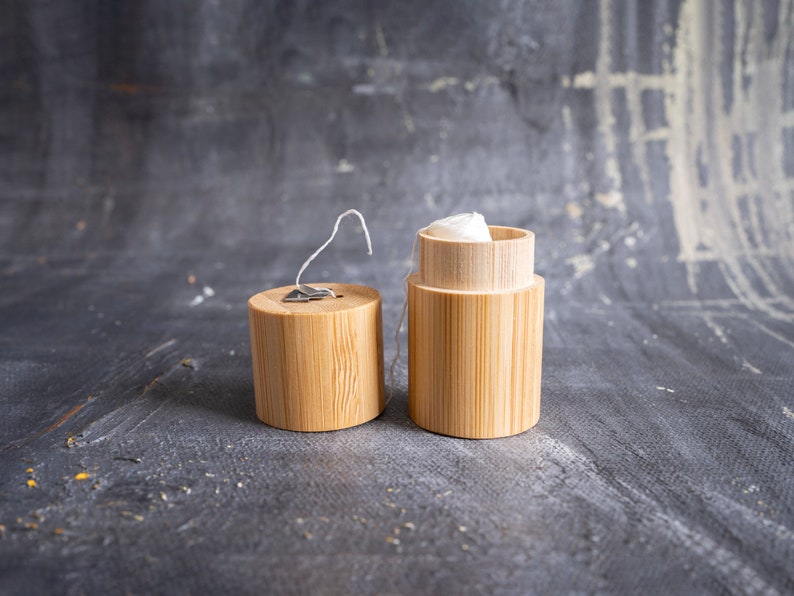 May include: A cylindrical bamboo dental floss container with a removable lid. The lid features a small metal cutter for the white floss, which is visible extending from the open container. The natural wood grain of the bamboo is clearly displayed.