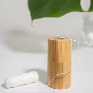 May include: A cylindrical bamboo dental floss container with a metal dispensing lid and a length of white dental floss extending from the top, accompanied by a separate small, coiled spool of white dental floss resting nearby.