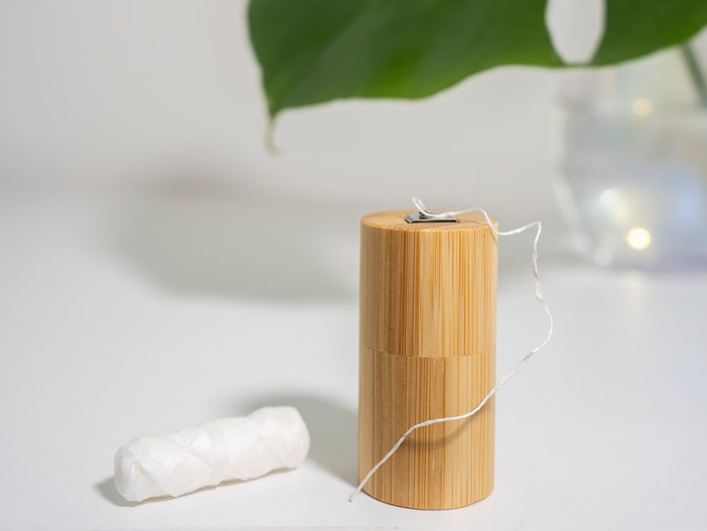May include: A cylindrical bamboo dental floss container with a metal dispensing lid and a length of white dental floss extending from the top, accompanied by a separate small, coiled spool of white dental floss resting nearby.