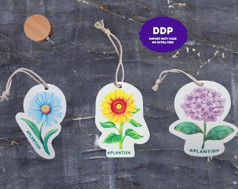 May include: Three hanging air fresheners featuring illustrations of a blue daisy, a yellow sunflower, and a purple hydrangea. Each white cutout shape includes a string loop and the green text PLANTISH. A small wooden disc is also included.
