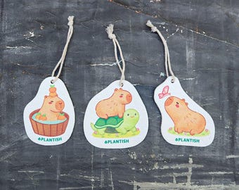 May include: Three white, ceramic ornaments with a capybara design. The first ornament features a capybara taking a bath with oranges. The second ornament features a capybara riding a turtle. The third ornament features a capybara with a butterfly flying above it. All ornaments have the word "Plantish" printed on them.