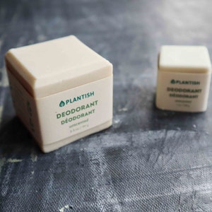 Puede incluir: Dos barras de desodorante beige en forma de cubo con etiquetas de papel color crema. La barra m&aacute;s grande tiene la etiqueta "PLANTISH DEODORANT D&Eacute;ODORANT unscented 99 g" y la m&aacute;s peque&ntilde;a "PLANTISH DEODORANT D&Eacute;ODORANT 28 g".