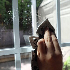 May include: A light-colored cleaning cloth with a black botanical print pattern being used to wipe a white-framed window pane. The cloth features a textured surface and is shown in motion, leaving a semi-circular streak of cleaning solution.