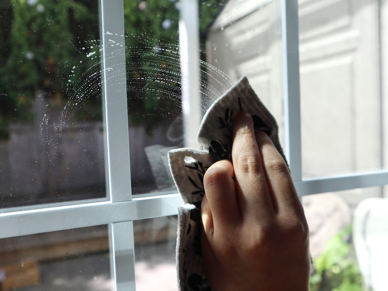 May include: A light-colored cleaning cloth with a black botanical print pattern being used to wipe a white-framed window pane. The cloth features a textured surface and is shown in motion, leaving a semi-circular streak of cleaning solution.