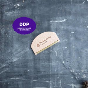 Peut inclure: Un outil anti-peluches en bois en forme de demi-cercle avec un bord métallique et le texte PLANTISH FUTURE accompagné d'une petite icône de feuille imprimé sur la surface en bois clair. L'outil est placé sur un fond gris texturé.