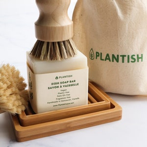 Pode incluir: Uma barra de sab&atilde;o para lavar lou&ccedil;a branca com um logotipo de folha verde e o texto "PLANTISH DISH SOAP BAR SAVON &Agrave; VAISSELLE" no r&oacute;tulo. A barra de sab&atilde;o est&aacute; em um suporte de sab&atilde;o para lavar lou&ccedil;a de bambu com uma escova de madeira.