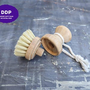 May include: A two-piece wooden dish brush with a detachable head featuring light-colored stiff bristles and a threaded screw mechanism. The handle has a concave shape and includes a white cotton rope loop attached to the side for hanging.
