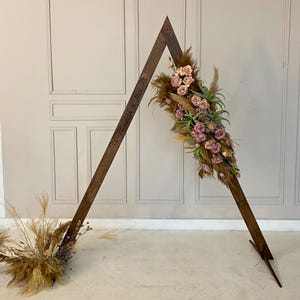 Könnte beinhalten: A-förmiger Hochzeitsbogen aus Holz, verziert mit getrocknetem Pampasgras, rosa Rosen und Grünzeug. Auf dem Bogen sind die Namen "Elisabeth & Max" eingraviert. Der Bogen steht auf einem weißen Boden vor einer neutralen Wand.