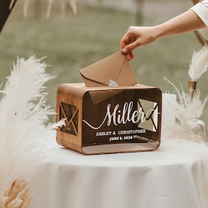 May include: A wooden wedding card box with the name "Miller" and the date "June 6, 2026" in white script. The box has a slot for cards and a clear front panel. A hand is placing a brown envelope into the box.