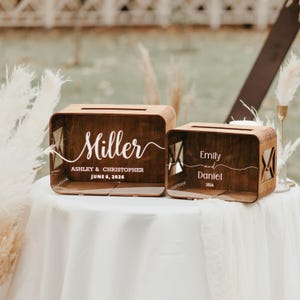 May include: Two rectangular dark brown wooden boxes with slots on top. The larger box features white text reading Miller, ASHLEY & CHRISTOPHER, JUNE 6, 2026. The smaller box features white text reading Emily and Daniel, 2026. Both have side cutouts.