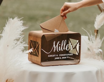 May include: A wooden wedding card box with the name "Miller" and the date "June 6, 2026" in white script. The box has a slot for cards and a clear front panel. A hand is placing a brown envelope into the box.