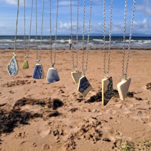 Könnte beinhalten: Eine Auswahl an Halsketten mit Anhängern aus Keramikscherben. Die Anhänger in verschiedenen Formen zeigen blau-weiße Muster und sind an Silberketten befestigt. Die Halsketten werden an einem Strand mit Meerblick im Hintergrund präsentiert.