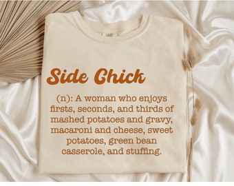 May include: A cream-coloured t-shirt featuring the words "Side Chick" in a brown, retro font. Underneath, a definition humorously describes a woman who enjoys mashed potatoes, macaroni and cheese, and other comfort foods. The shirt is displayed on a white satin surface.