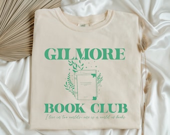 Może przedstawiać: Beżowa koszulka z zielonym nadrukiem, na którym widnieje napis "GILMORE BOOK CLUB" oraz "Żyję w dwóch światach: jeden to świat książek".
