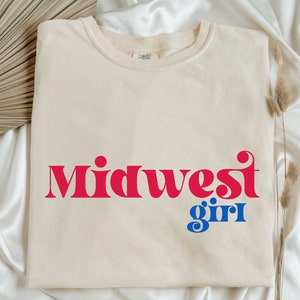Puede incluir: Una camiseta de color crema con el texto "Midwest girl" impreso en rojo y azul. El texto está en un estilo de fuente retro.