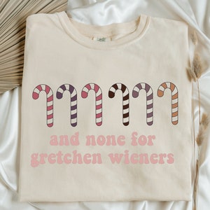 May include: A beige t-shirt with a graphic of candy canes and the text "and none for gretchen wieners".