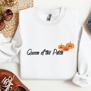 Könnte beinhalten: Weißes Sweatshirt mit dem Text "Queen of the Patch" in schwarzer Schrift, begleitet von drei orangefarbenen Kürbissen. Auf den Kürbissen stehen die Namen "Jane", "Freddie" und "Chucky". Herbstliches Thema.