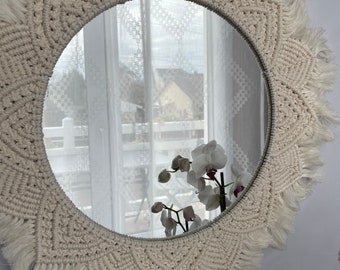 Peut inclure: Miroir rond en macramé crème, avec une bordure à franges. Le cadre présente un motif géométrique. Le miroir reflète une fenêtre avec des rideaux blancs, une maison et une orchidée blanche.