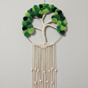 Puede incluir: Un tapiz de pared de macramé hecho a mano con un diseño de árbol. El árbol tiene un tronco y ramas de color crema, con una corona de pompones verdes y negros. El diseño está en un anillo dorado, con hebras de macramé color crema y cuentas de madera.
