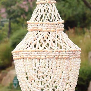 Peut inclure: Un lustre suspendu fait de coquillages naturels. Le lustre a une forme conique avec plusieurs niveaux, chacun orné de petits coquillages crème. Le design crée un luminaire décoratif texturé, parfait pour la décoration côtière.