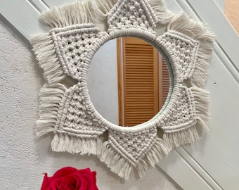 Peut inclure: Un miroir rond avec un cadre en macramé crème. Le cadre présente un motif en étoile avec des éléments triangulaires et à franges. Le miroir est accroché à un mur blanc, avec une rose rouge au premier plan.