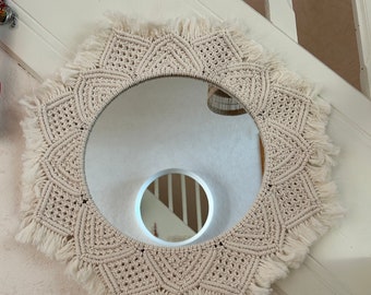 Peut inclure: Miroir rond avec un cadre en macramé crème. Le macramé présente des motifs géométriques et des franges. Le miroir est fixé sur un mur blanc. L'ensemble est de style bohème et artisanal. Le diamètre est d'environ 30 cm.