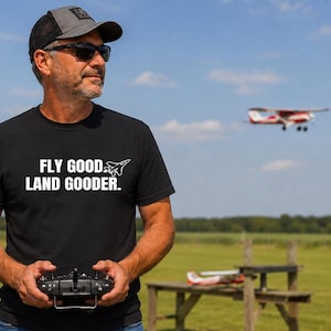May include: A black short-sleeved t-shirt featuring the white text FLY GOOD LAND GOODER and a small graphic of a fighter jet. The shirt is made of a solid black fabric with a crew neckline and a standard fit, displaying the printed design on the chest.