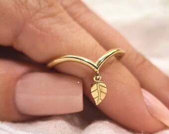 Puede incluir: Un anillo dorado con un diseño en forma de V y un pequeño dije de hoja. El anillo se muestra en un dedo, mostrando sus delicados detalles y su acabado pulido. El dije de hoja cuelga elegantemente del centro del anillo.