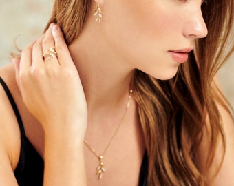 May include: A woman wearing a black top with a gold necklace and earrings featuring a delicate leaf design. The necklace and earrings are both made of gold and have a simple, elegant style.