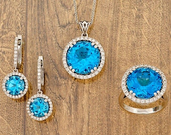 May include: A set of blue topaz jewelry featuring a pendant necklace, a pair of earrings, and a ring. Each piece has a round blue topaz stone surrounded by a halo of smaller diamonds.