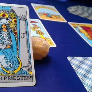 May include: The High Priestess tarot card features a woman in a blue robe sitting on a throne with a crescent moon and a book in her lap. The card is surrounded by other tarot cards on a blue surface.
