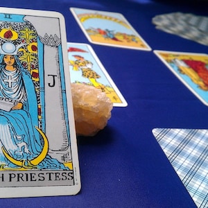May include: The High Priestess tarot card features a woman in a blue robe sitting on a throne with a crescent moon on each side. The card is part of a tarot card spread on a blue surface.